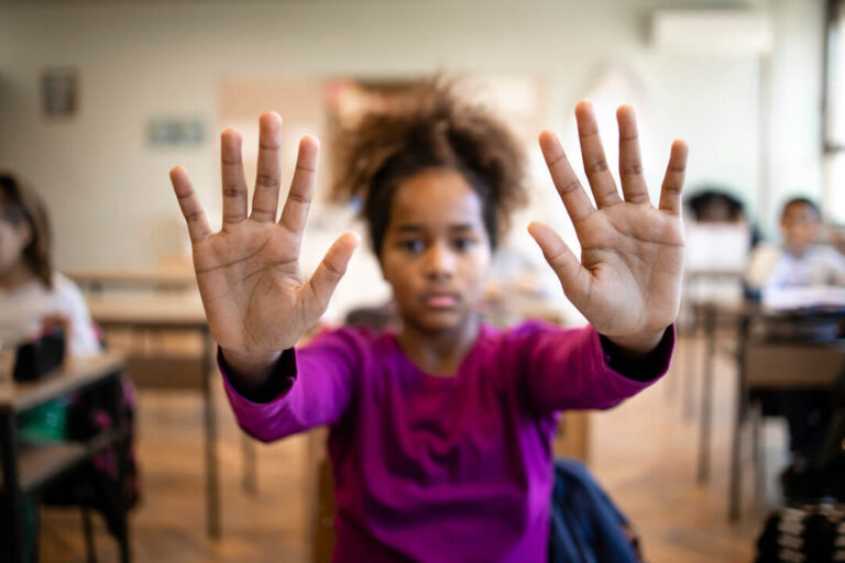 Educação antirracista: imagem mostra menina em sala de aula com mãos esticadas para frente.